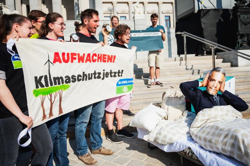 Aktivist:innen halten Banner in der Hand, worauf steht: "Aufwachen: Klimaschutz jetzt!". Ein Aktivist mit einer Nehammer-Maske liegt daneben im Bett und hält sich die Ohren zu.