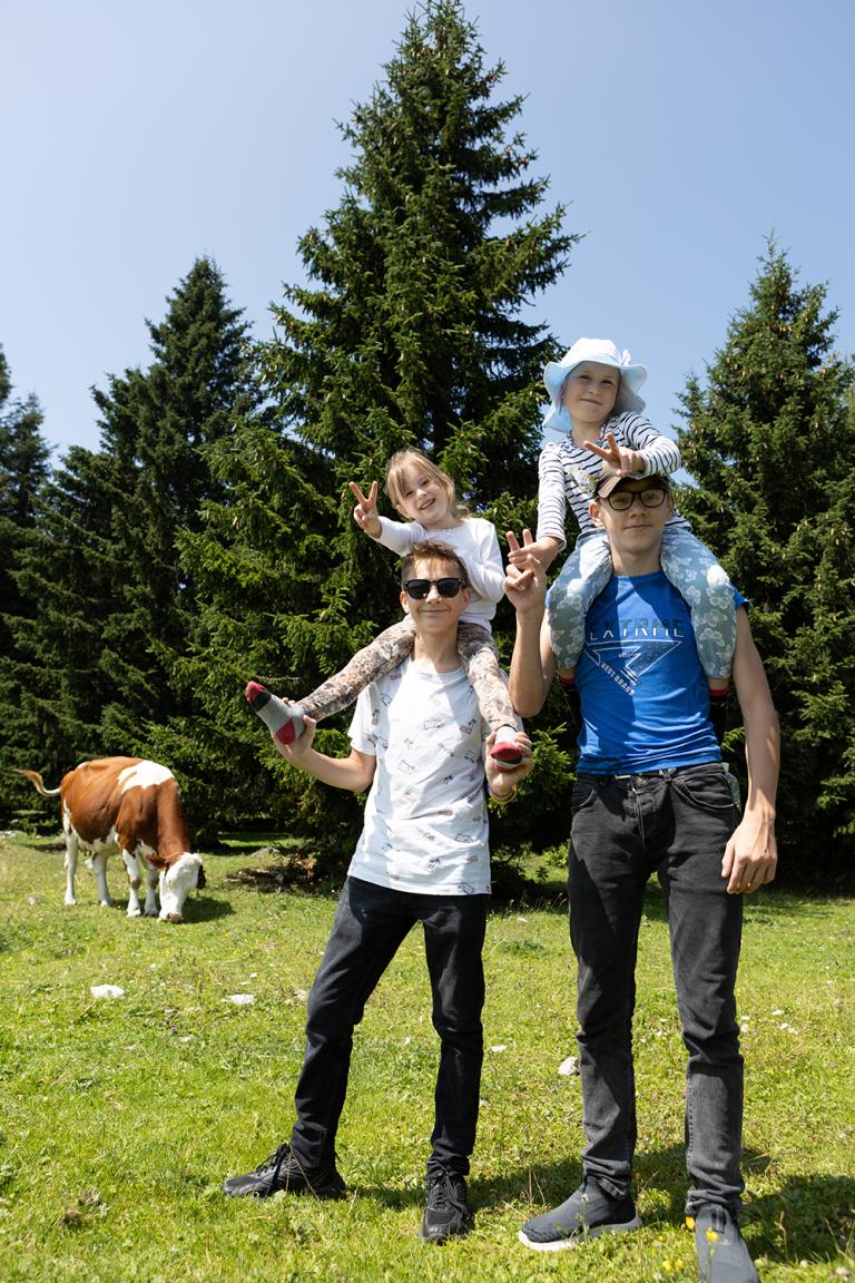 zwei Kinder sitzen auf den Schultern von zwei Erwachsenen, im Hintergrund sieht man eine Kuh