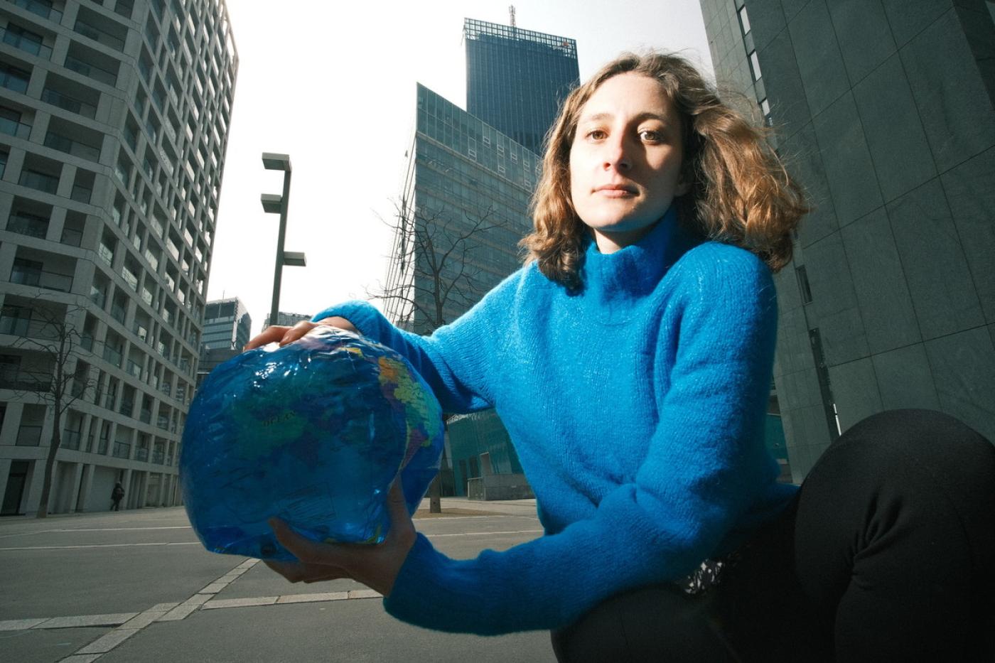 Anna Leitner trägt einen blauen Pullover, sie kniet am Boden vor hohen Bürogebäude und hält eine Plastik-Weltkugel in der Hand