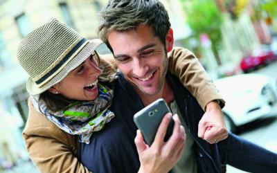 Eine Frau mit Hut umarmt einen Mann und beide schauen freudig in ein Smartphone.