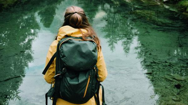 Frau mit gelber Regenjacke und Rucksack steht mit dem Rücken zur Kamera und schaut auf einen See, dahinter befinden sich die Berge im Nebel
