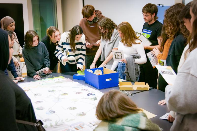 Schülergruppe steht um einen Tisch und sie schauen interessiert in eine blaue Box. Einige von ihnen halten eine Karte in den Händen. 