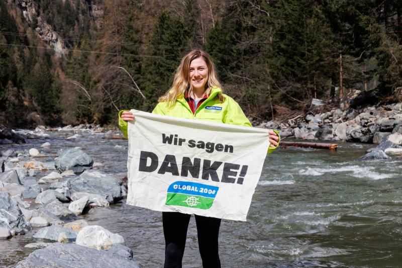 Viktoria Auer steht neben einem Fluss. In der Hand hält sie einen Banner mit der Aufschrift "Wir sagen Danke"