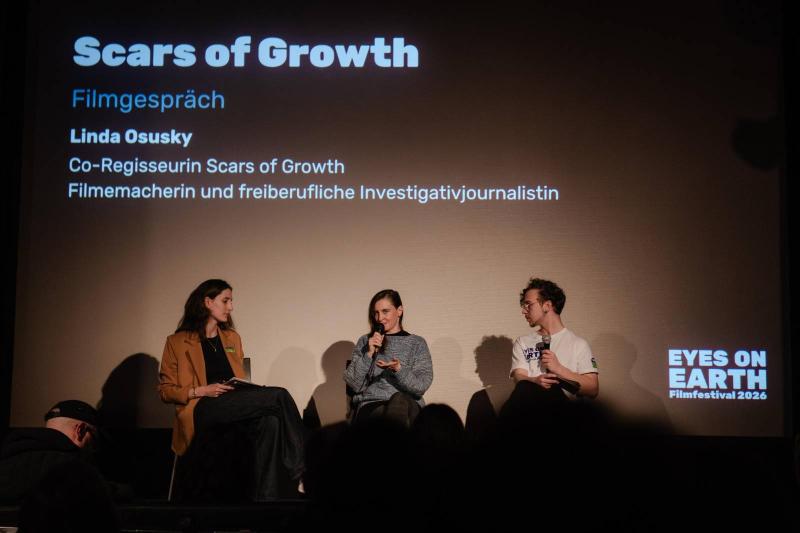Blick auf die Bühne eines Kinosaals, auf der 3 Personen sitzen, eine davon spricht in ein Mikro, dahinter sieht man "Scars of Growth" an die Wand gebeamt