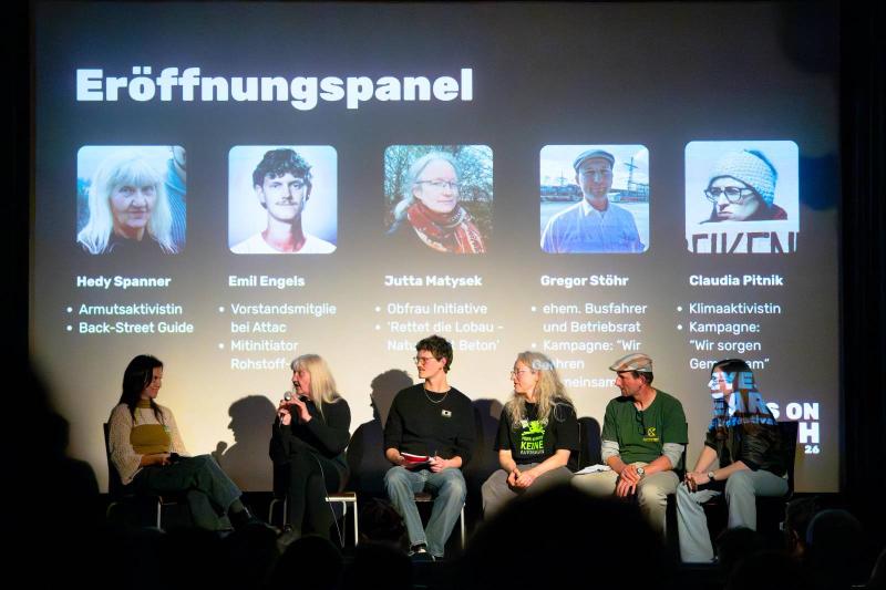 Sechs Personen sitzen auf einer Bühne im Kino und diskutieren, dahinter ist groß Eröffnungspanel mit Infos hingebeamt