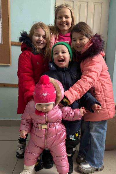 4 lachende Kinder halten 1 Kleinkind in rosa Skianzug fest