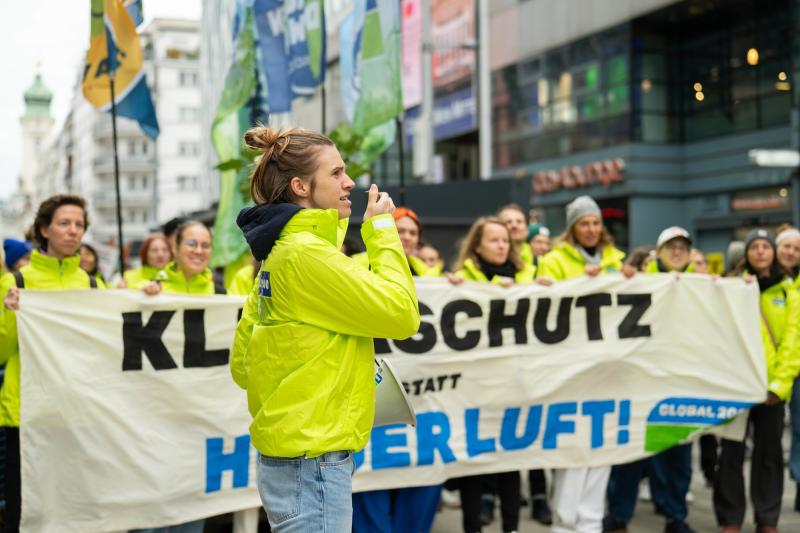 Demonstrierende in GLOBAL 2000 Jacken, im Vordergrund steht eine Person mit Megafon, im Hintergrund sind mehrere Personen die ein Banner halten mit der Aufschrift "Klimaschutz statt heißer Luft"