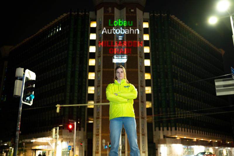 In der Mitte des Bildes steht eine junge Frau mit entschlossenem Blick und verschränkten Armen vor einem Gebäude, dem österreichischen Verkehrsministerium. Die Frau trägt eine Jacke mit einem Global 2000 Logo. Auf das Gebäude hinter ihr ist ein Schriftzug gebeamt "Lobau Autobahn = Milliardengrab!"