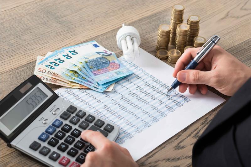 Auf einem Tisch liegen ein Taschenrechner, Geld, eine Glühbirne und ein Zettel mit einer Tabelle und man sieht zwei Hände, die schreiben und auf den Taschenrechner tippen