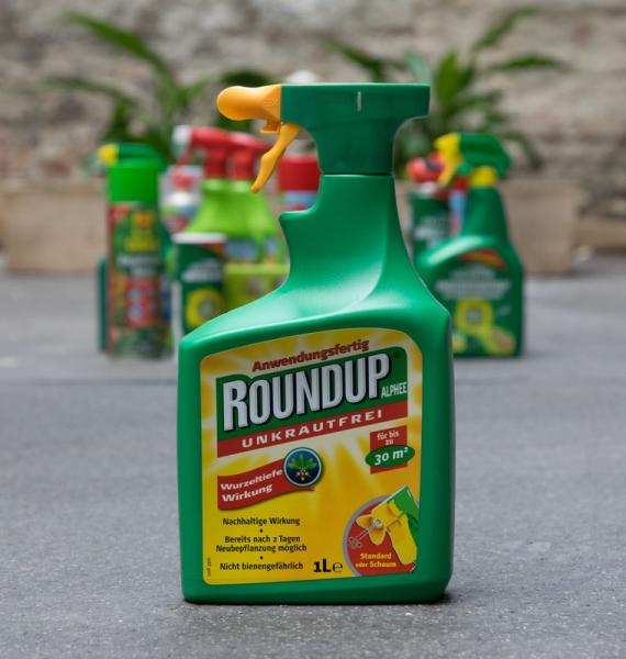 Grüne Sprühflasche mit gelbem Sprühkopf und Etikett 'ROUNDUP UNKRAUTFREI' im Vordergrund, unscharfe weitere Flaschen im Hintergrund