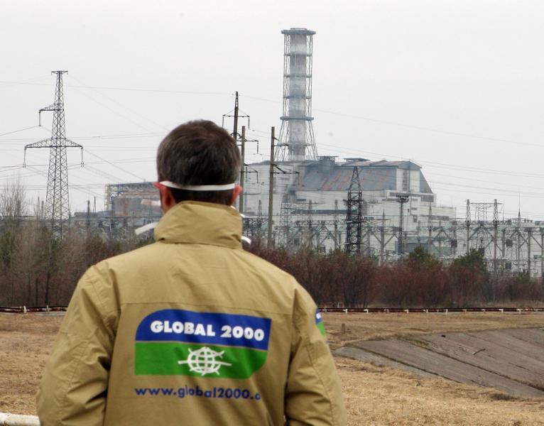 Person mit beiger Jacke und Aufschrift "GLOBAL 2000" auf dem Rücken steht vor einem Industriegebäude mit hohem Schornstein und Stromleitungen