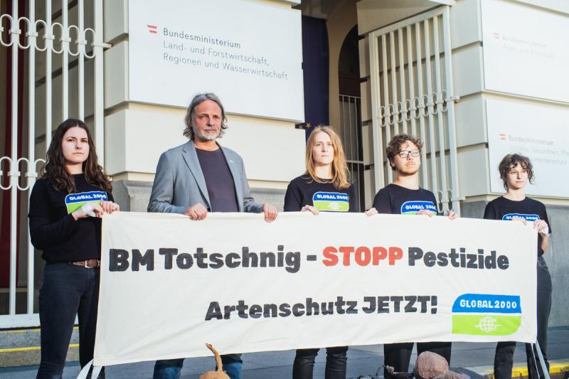 Fünf Personen halten vor einem Gebäude ein Banner mit der Aufschrift 'BM Totschnig - STOPP Pestizide Artenschutz JETZT'