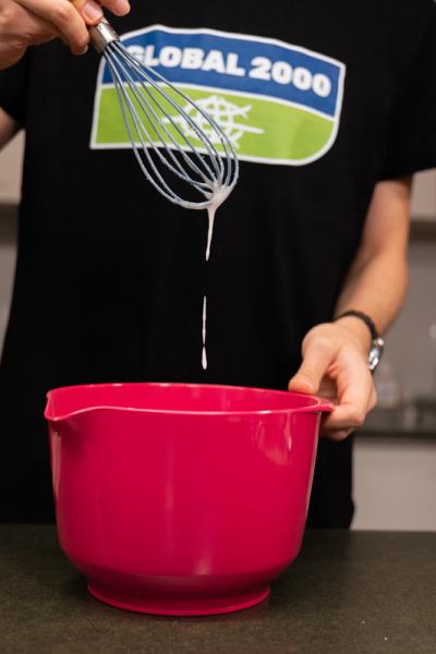 Person mit schwarzem Shirt und GLOBAL 2000 Logo hält einen Schneebesen mit tropfendem Teig über eine rote Rührschüssel in der Küche.