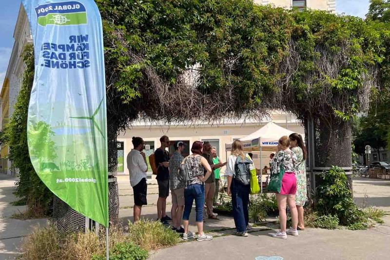Gruppe von Personen vor einem Infostand von GLOBAL 2000 mit Banner „Wir kämpfen für das Schöne.“