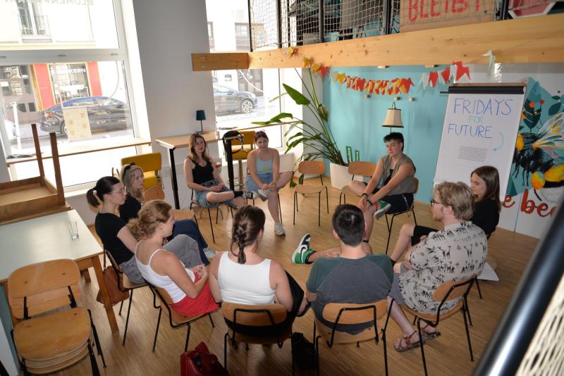 Junge Menschen sitzen in einem hellen Raum im Café-Stil im Kreis und nehmen an einem „World Café“-Dialog für Jugendliche teil; „Fridays for Future“-Poster und Girlanden.