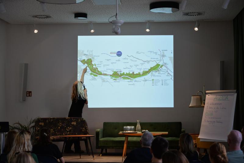 Jutta Matysek von Lobau bleibt! zeigt während eines Vortrags für das Jugenddialog-Event auf eine projizierte Karte des Gebiets Donau-Auen/Lobau, während die Teilnehmer zuschauen; daneben steht ein Flipchart mit der Tagesordnung.