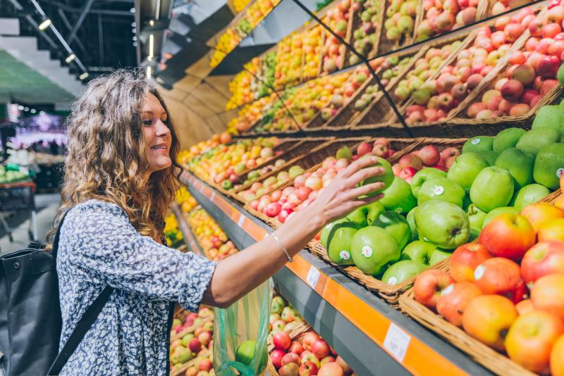Frau greift im Supermarkt in ein Obstregal und nimmt einen Apfel