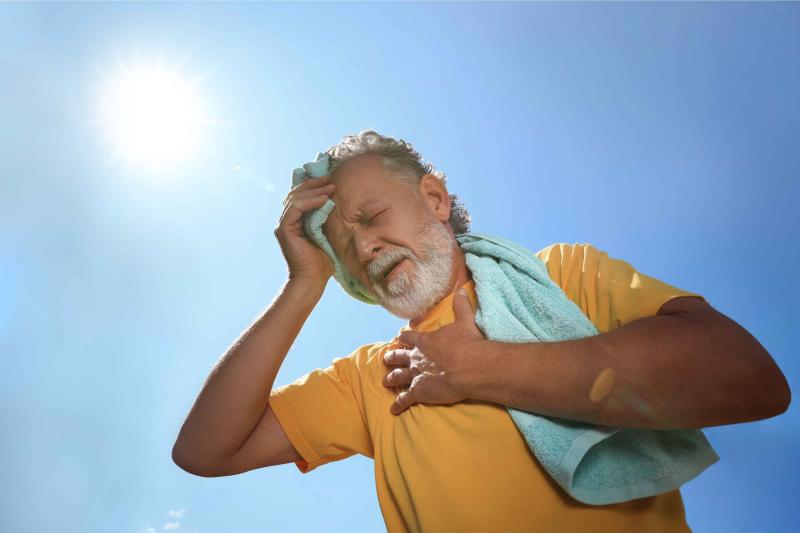 Älterer Mann in gelbem Shirt mit Handtuch greift sich mit schmerzverzerrtem Gesicht auf Kopf und Brustbereich, über ihm Sonne und blauer Himmel