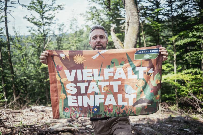 Biodiversitätsexperte Dominik Linhard hält im Wald eine Fahne mit der Aufschrift 'VIELFALT STATT EINFALT' und dem Logo 'GLOBAL 2000'