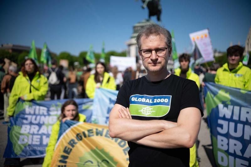 Johannes Wahlmüller steht mit schwarzem T-Shirt mit Aufdruck "GLOBAL 2000" vor einer Gruppe von Menschen mit bunten Bannern bei einer Demonstration im Freien