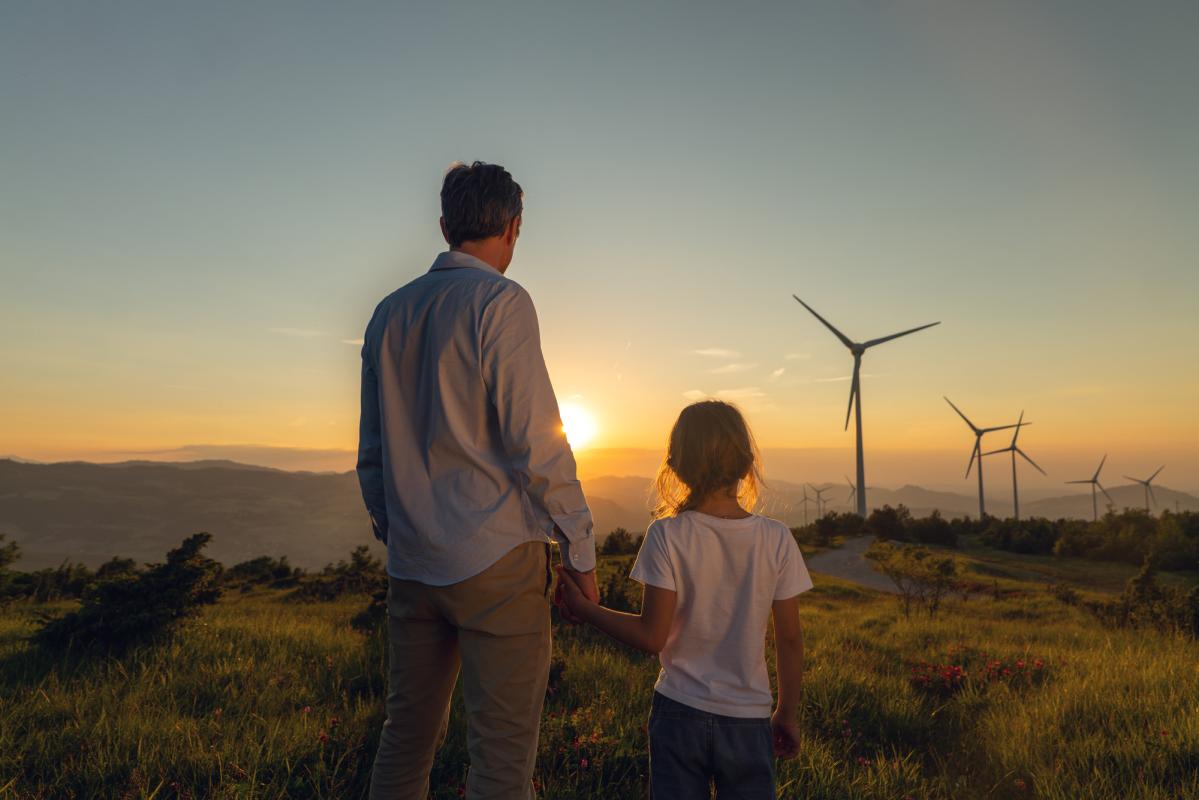 Vater und Kind blicken Richtung Sonnenaufgang in Landschaft mit Windrädern