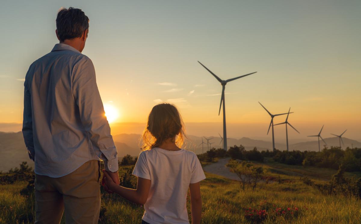 Vater und Kind blicken Richtung Sonnenaufgang in Landschaft mit Windrädern