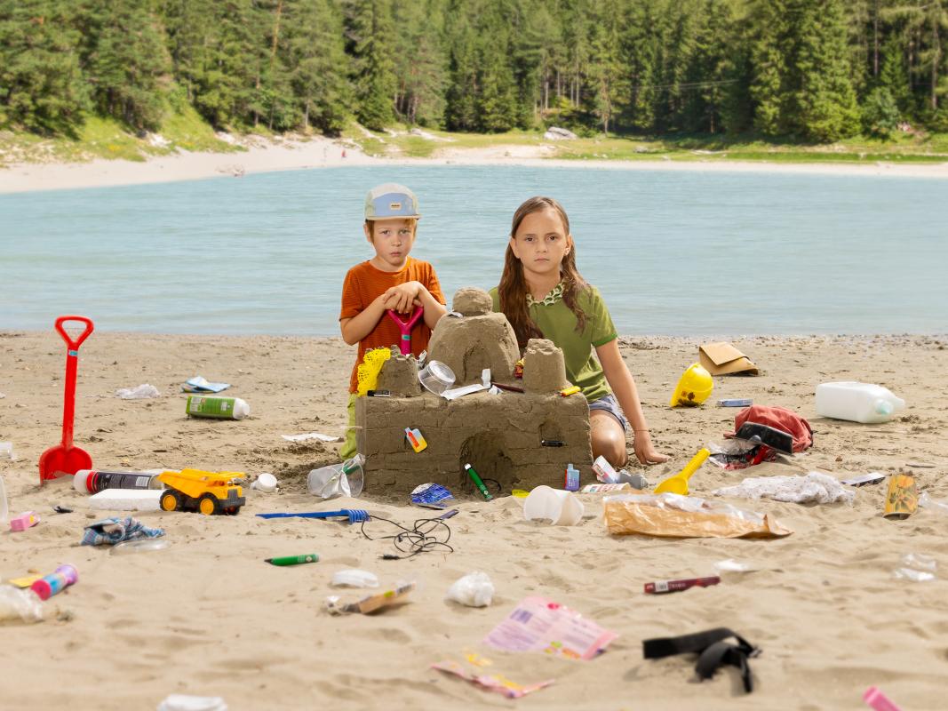 2 Kinder bauen eine Sandburg mit Müll vor österreichischer Landschaft