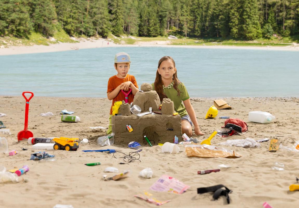 2 Kinder bauen eine Sandburg mit Müll vor österreichischer Landschaft