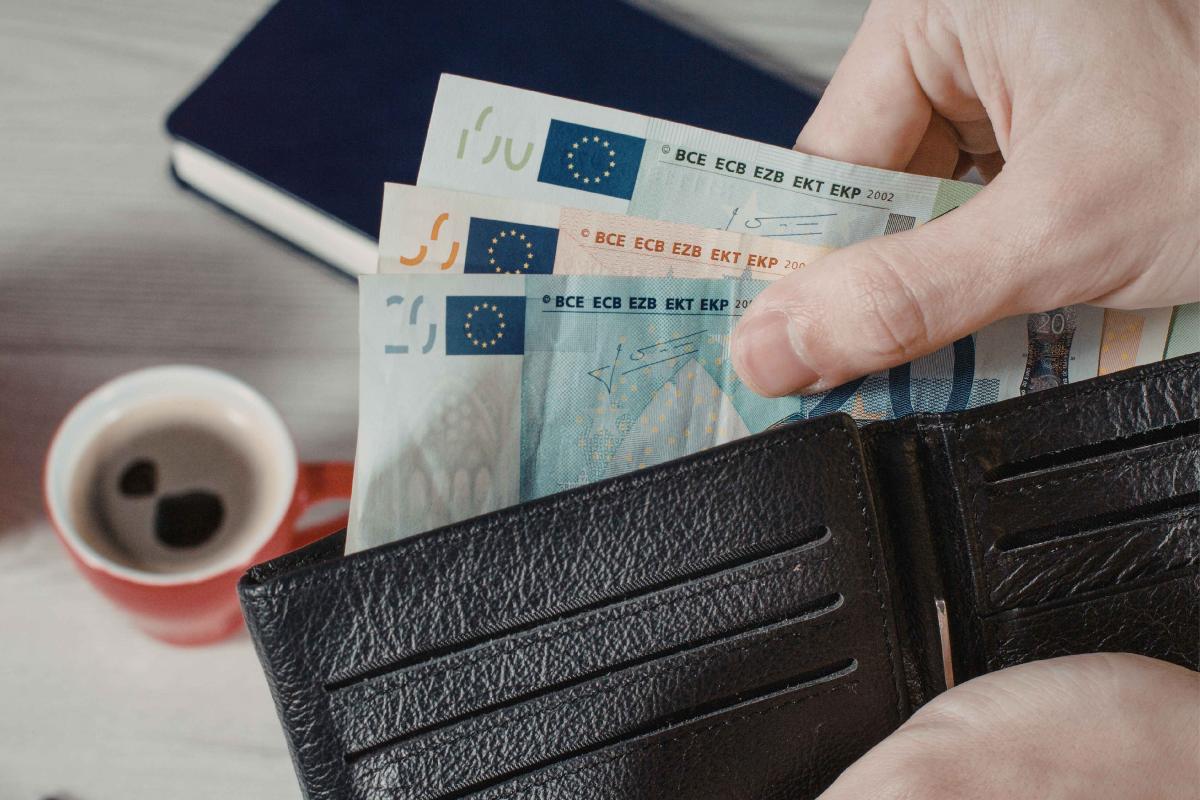 Eine Hand zieht mehrere Euro Geldscheine aus einer Geldbörse, auf dem Tisch dahinter steht ein Kaffee und es liegt ein Notizbuch daneben