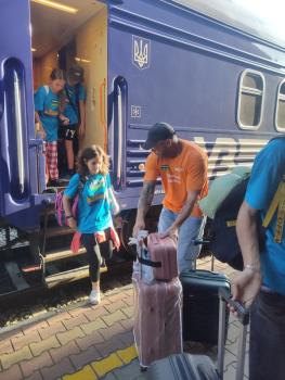 Kinder steigen aus einem blauen Zug mit ukrainischem Wappen aus, ein Mann hilft beim Tragen von Koffern auf einem Bahnsteig