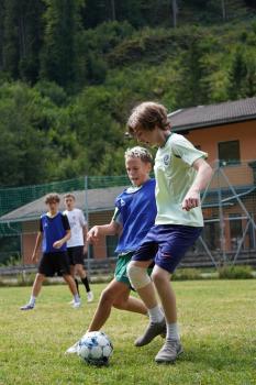 wei Jungen spielen Fußball auf einer Wiese, einer trägt ein blaues Trikot, der andere ein helles T-Shirt, im Hintergrund zwei weitere Jungen und ein Gebäude mit Zaun.