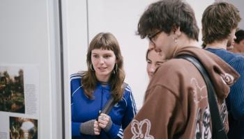 Drei junge Menschen stehen zusammen in einem Innenraum und betrachten Bilder oder Texte an einer Wand. Einer trägt eine blaue Jacke, ein anderer einen braunen Hoodie mit Mustern. Sie wirken vertieft im Gespräch, während sie die Ausstellung betrachten.