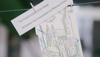 Eine handgezeichnete Skizze einer Kathedrale hängt an einer Schnur mit einer Wäscheklammer. Darüber ist ein Zettel auf Deutsch befestigt, auf dem steht: „Da der Stephansdom so riesig ist, da passen doch unendlich viele Pflanzen drauf!