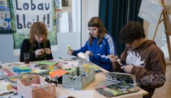 Drei Personen sitzen an einem Tisch, der mit Bastelmaterialien bedeckt ist, und fertigen Kunstwerke oder Plakate an. Bunte Papiere, Marker und Körbe liegen auf dem Tisch verstreut. Im Hintergrund ist ein Schild zu sehen, auf dem „Lobau bleibt!“ steht.
