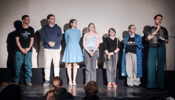 7 Personen stehen auf einer Bühne, die Person ganz rechts spricht in ein Mikrophon