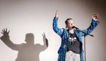 Frau auf Bühne vor Mikrophon spircht ins Mikro mit erhobenen Armen