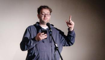 Mann vor Mikrophon auf einer Bühne sieht auf sein Handy und erklärt etwas mit erhobenem Zeigefinger