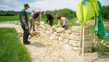 Menschgen bauen eine Trockensteinmauer, davor hängt eine hellgrüne Jacke mit der Aufschrift GLOBAL 2000