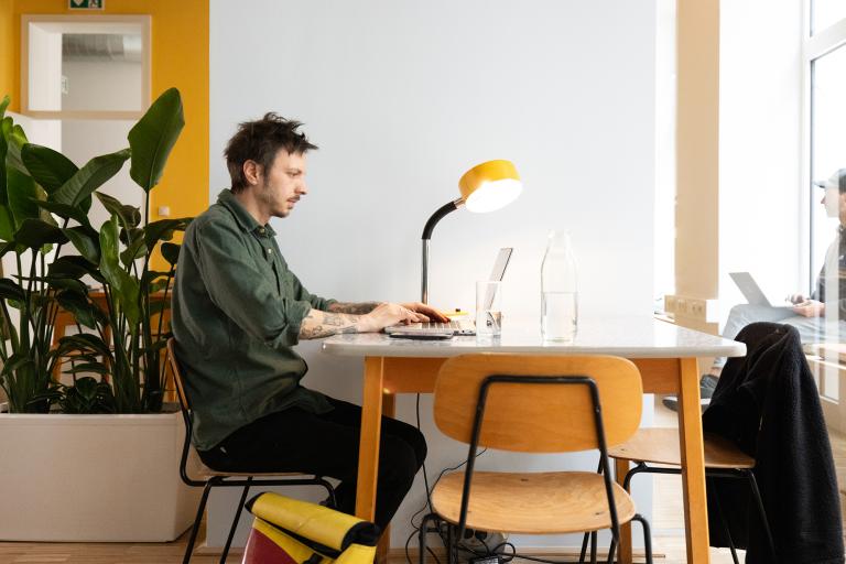 Ein Mann arbeitet an einem Tisch an seinem Laptop. Eine gelbe Lampe steht danben, vor einer blauen Wand.