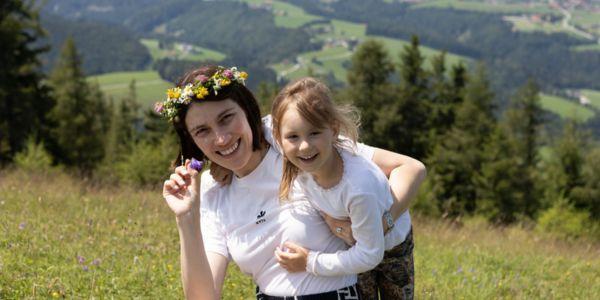 Eine Frau in den Bergen mit einem Blumenkranz im Haar hält ein blondes, lachenes Mädchen im Arm