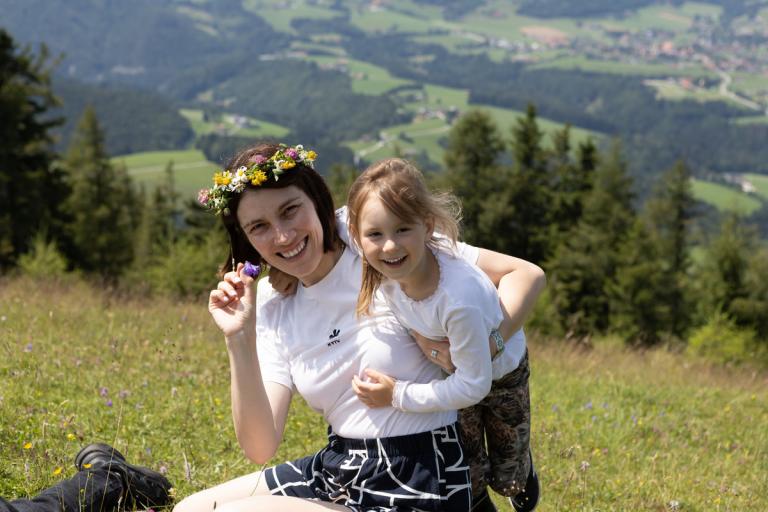 Eine braunhaarige Frau sitzt auf einer Wiesen in den Bergen mit einem Blumenkranz auf dem Kopf und hält ein blondes, lachendes Kind hoch