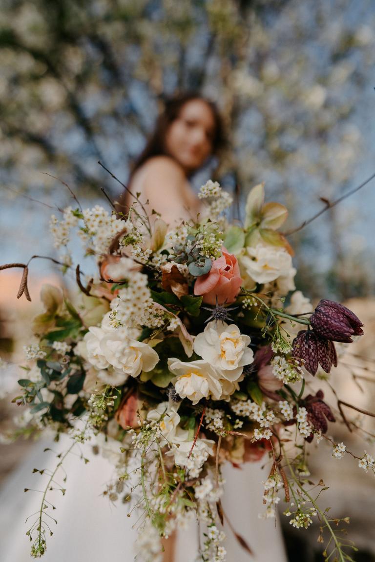 Blumenstrauß wird von einer jungen Frau in einem weißen Kleid in die Kamera gehalten. Der Hintergrund ist verschwommen. 