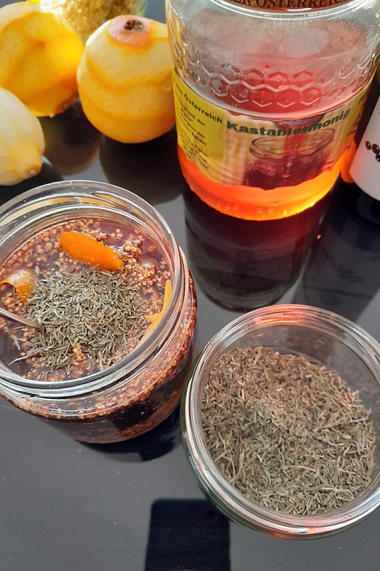 Drei Gläser aus der Vogelpersektive: in einem ist Honig, im anderen Oxymel und im dritten sind Kräuter. Daneben liegen Zitronen. 