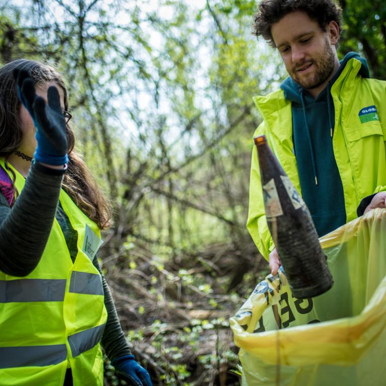 Ein Mann mit GLOBAL 2000 Jacke hält einen Müllsack auf. Ein Mädchen wirft eine Getränkeflasche hinein. 