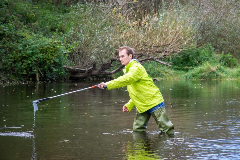 Ein Mann mit GLOBAL 2000 Jacke steht im Fluss und holt eine Wasserprobe