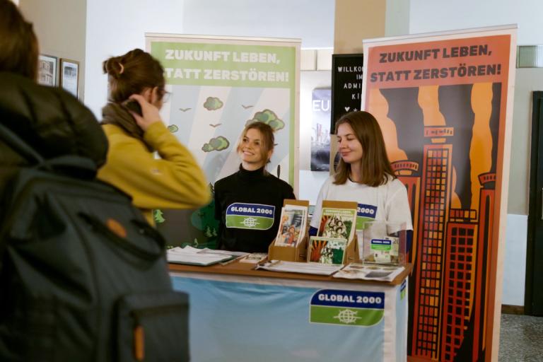 Zwei ehrenamtliche Mitarbeiterinnen stehen bei einem Info-Stand von GLOBAL 2000 und reden mit einer interessierten Person