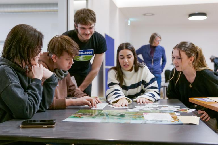 Ein Mann mit einem GLOBAL 2000 Shirt beugt sich über einen Tisch und hört Schüler:innen zu, wie sie ihm eine Frage stellen.
