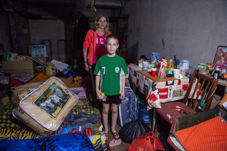 Junge und Mutter stehen in einem dunklen Zimmer, neben ihnen befinden sich Hilfspenden