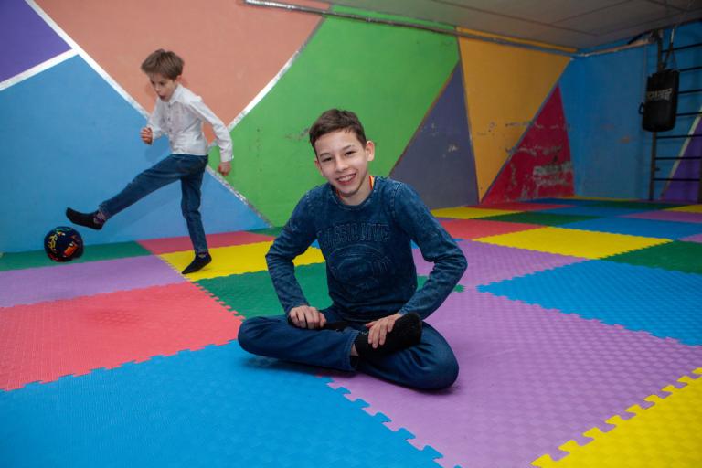 Bus sitzt in einen Schutzbunker und lacht in die Kamera, hinter ihm spielt ein Kind Fußball
