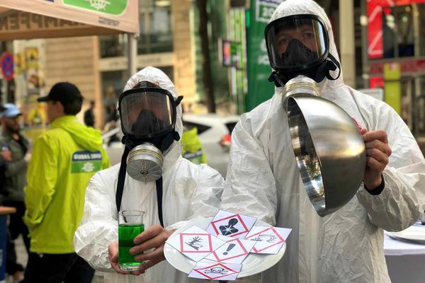 Zwei Personen in weißen Schutzanzügen und Atemschutzmasken halten ein Glas mit grünem Flüssigkeit und ein rundes Metallobjekt, davor ein Papier mit Gefahrensymbolen.